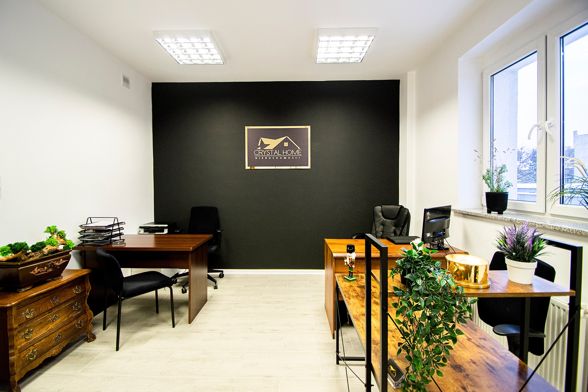Jasne biuro nieruchomości z dwoma biurkami, czarną ścianą z logo Crystal Home, roślinami doniczkowymi na parapecie i drewnianą komodą z bonsai.