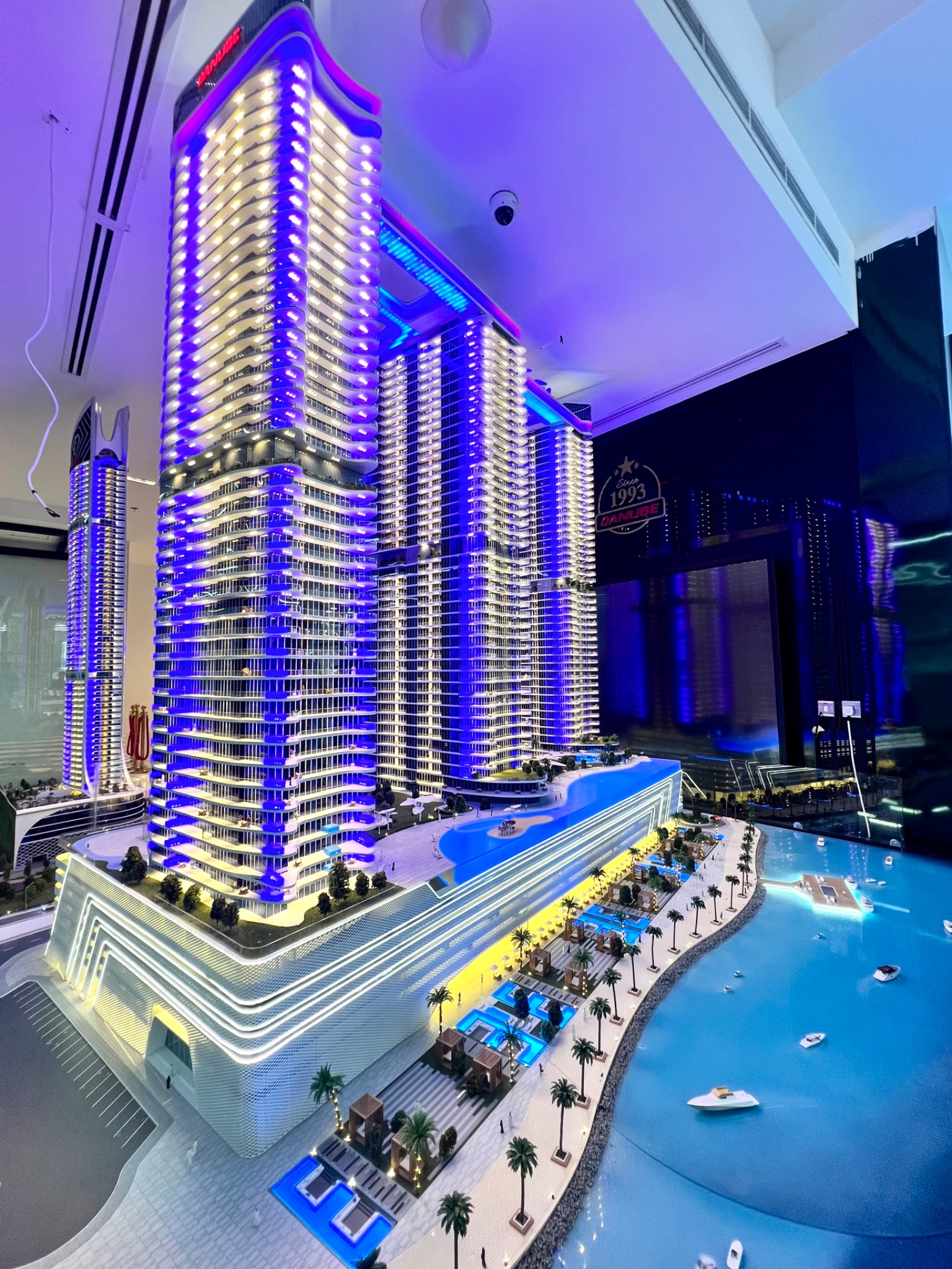 Architektoniczna makieta kompleksu budynków z basenami i palmami, podświetlona na niebiesko i biało, z widocznym logo firmy Danube z 1993 roku w tle.
