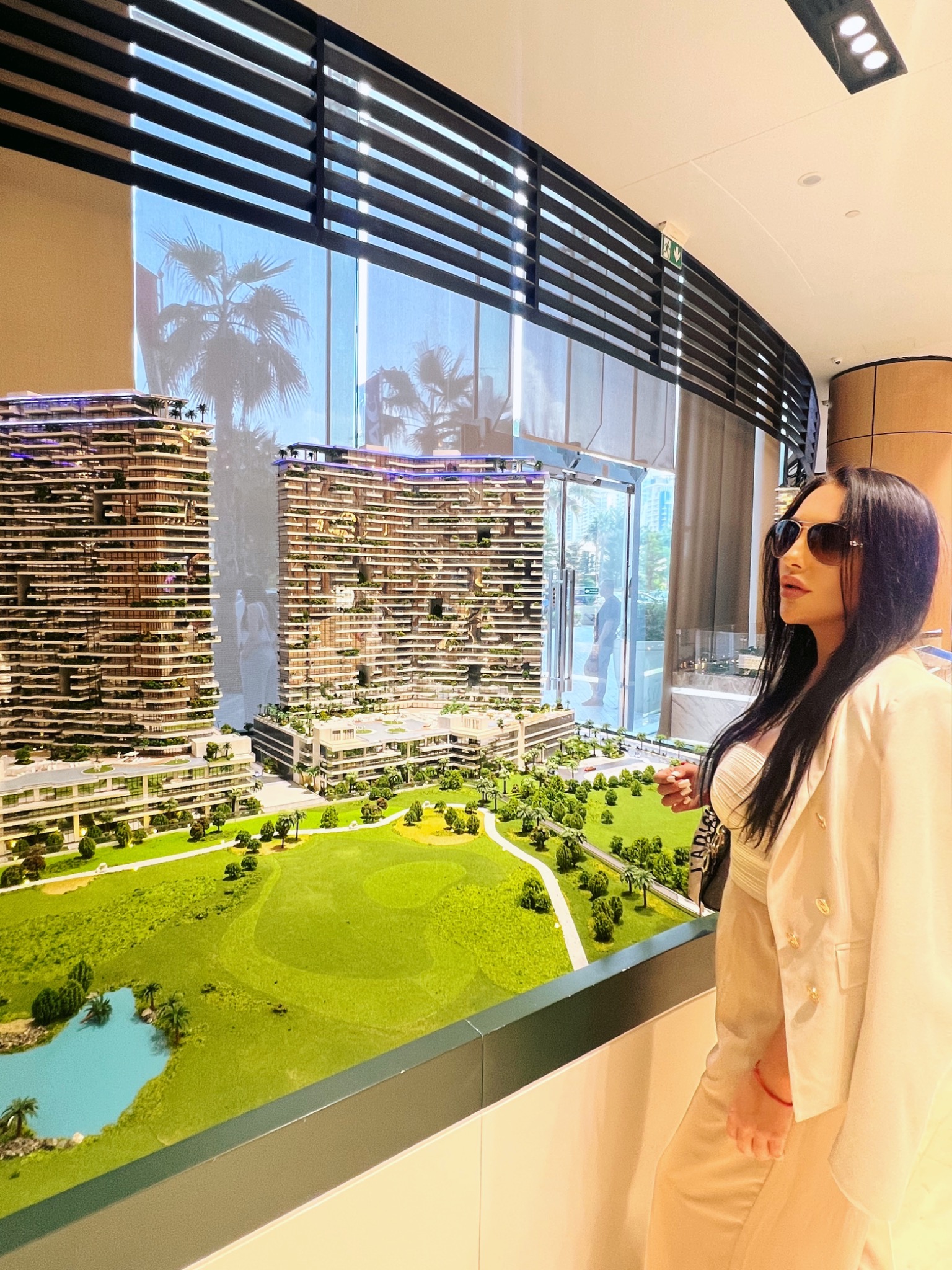 Kobieta w okularach przeciwsłonecznych ogląda makietę osiedla z budynkami wielorodzinnymi i polem golfowym, widoczne palmy na tle okna.