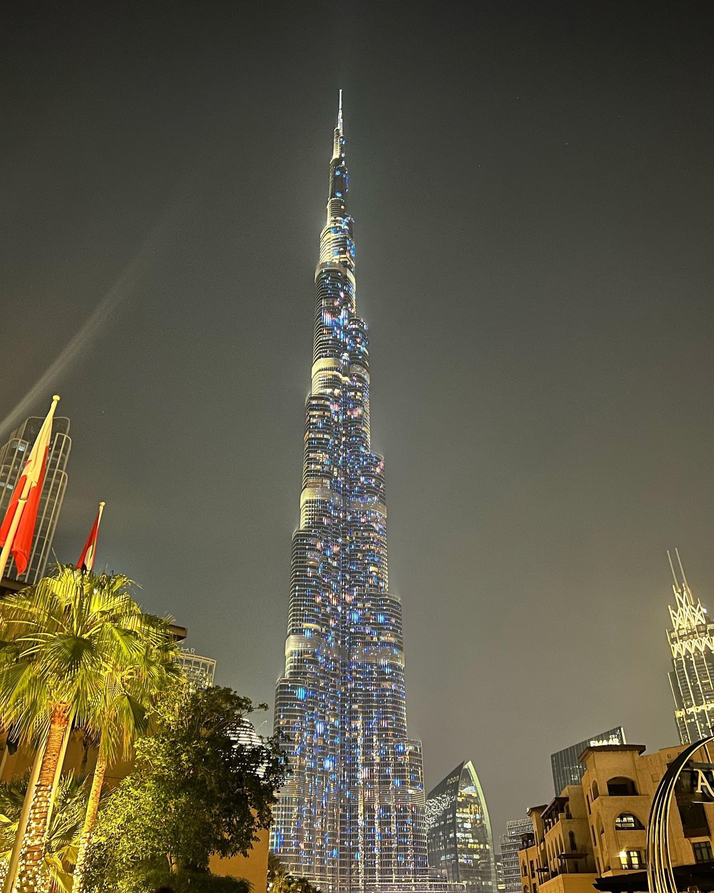 Nocne ujęcie drapacza chmur Burj Khalifa w Dubaju, z flagami i palmami na pierwszym planie, oświetlonego niebieskimi i białymi światłami.