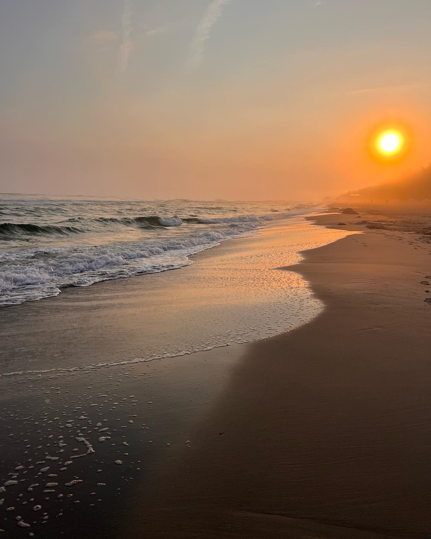 Morski brzeg o zmierzchu: fale delikatnie obmywają piasek, a słońce tonie w pomarańczowej mgle.