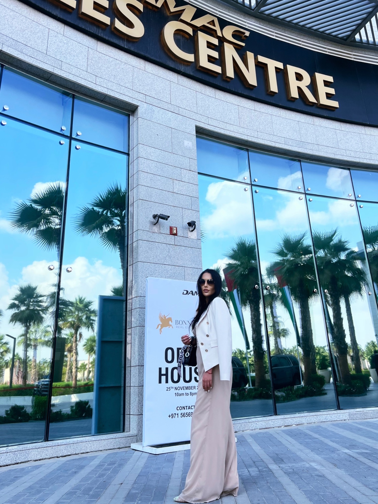 Kobieta w jasnym stroju stoi przed budynkiem 'MAC CENTRE' z dużymi oknami, w których odbijają się palmy i niebo, obok tablica informacyjna 'BONN REAL ESTATE OPEN HOUSE 25th NOVEMBER'