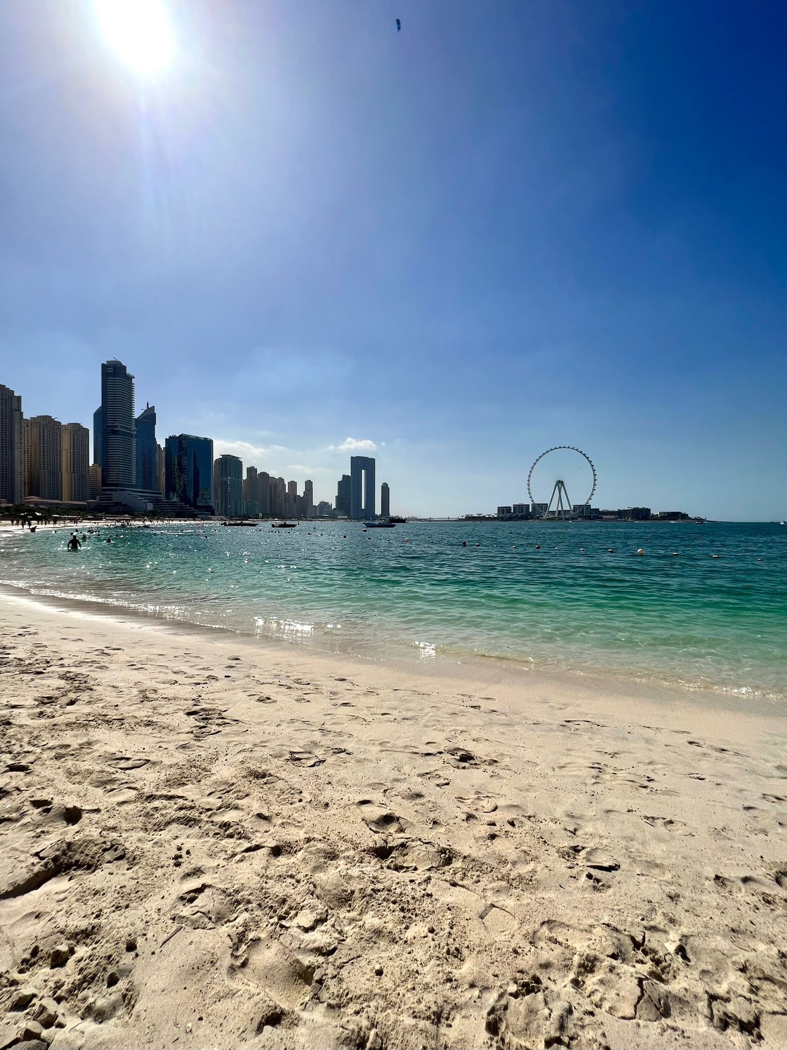 Słoneczny widok na plażę z piaskiem w odcieniach beżu, turkusową wodą, panoramą wieżowców w tle i diabelskim młynem na horyzoncie.