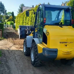 Ż&oacute;łta ładowarka Kramer Allrad 850 w trakcie pracy, widoczne ślady opon na piaszczystym podłożu, z tyłu usypisko ziemi i zielony żywopłot.