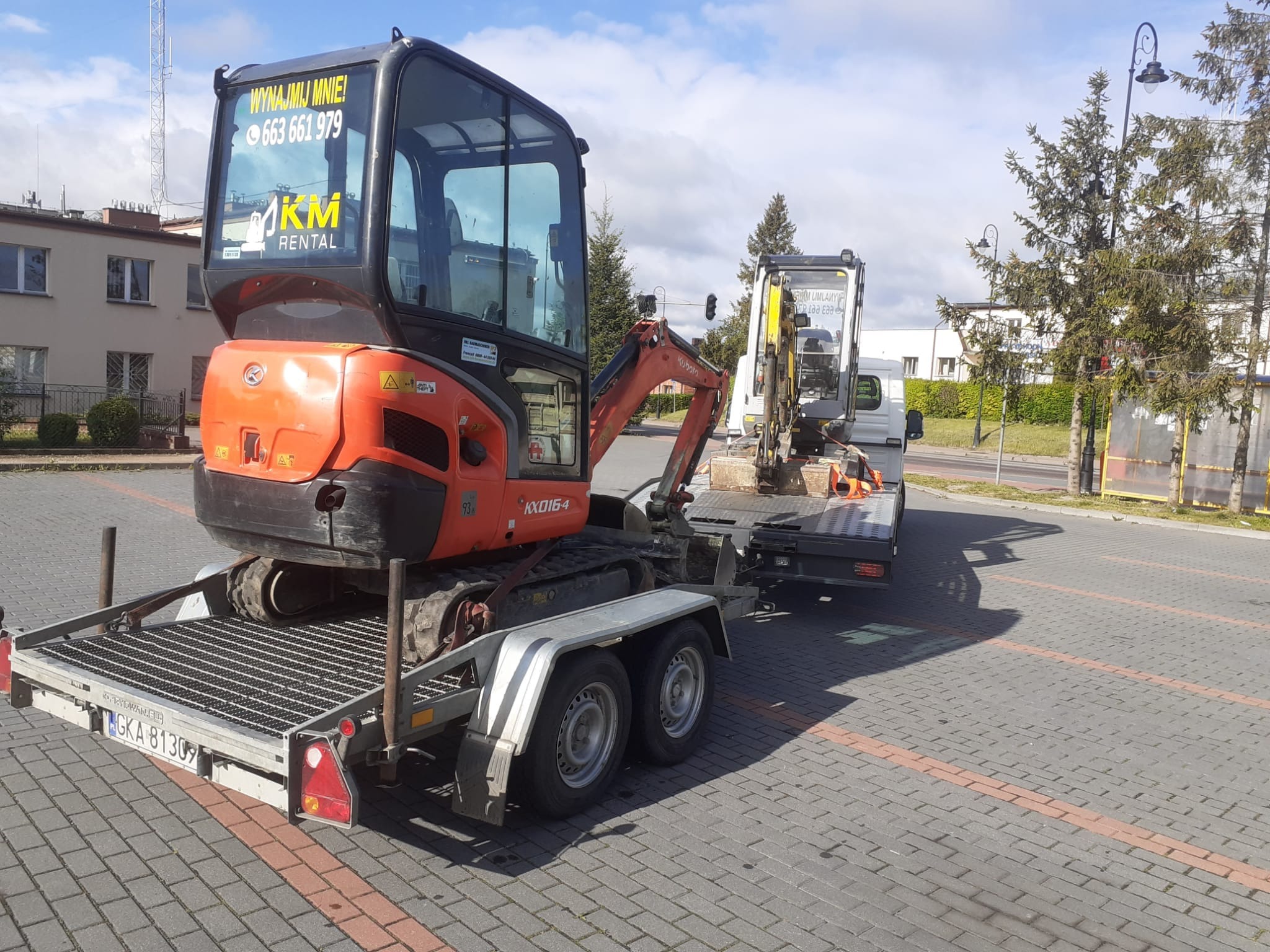 Pomarańczowa mini koparka Kubota KX016-4 z naklejką 'Wynajmij mnie!' na szybie, przewożona na przyczepie w Garczu. W tle zaparkowana biała furgonetka i budynki.