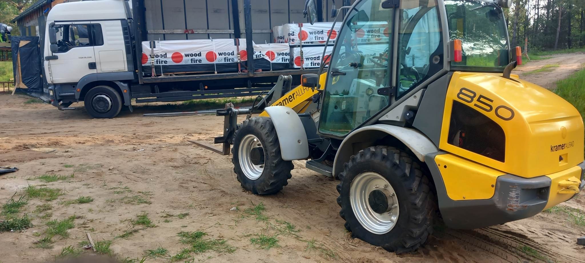 Żółto-szara ładowarka teleskopowa Kramer 850 z widłami, stojąca na piaszczystym terenie obok ciężarówki z ładunkiem drewna opałowego w opakowaniach z napisem 'Wood of Fire'.