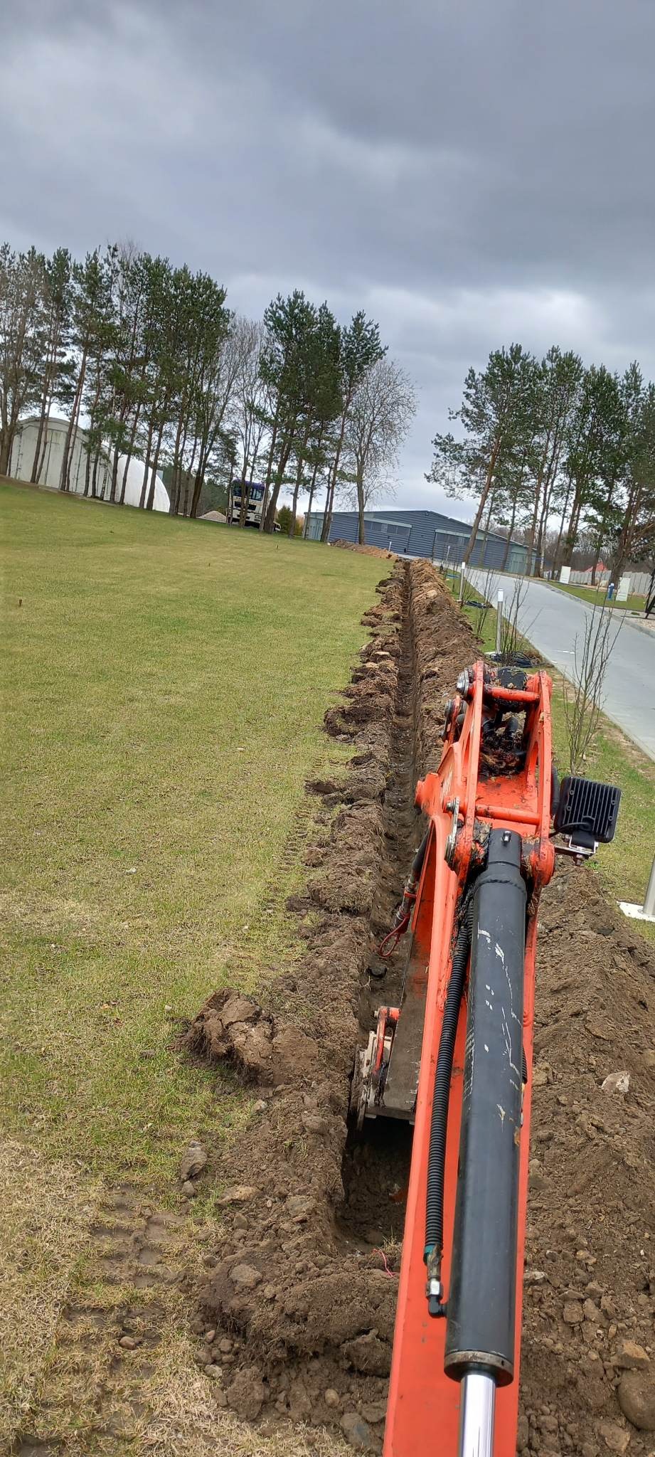 Pomarańczowa koparka wykonuje wąski wykop wzdłuż trawnika, widoczne ślady opon, w tle budynek i drzewa pod pochmurnym niebem.