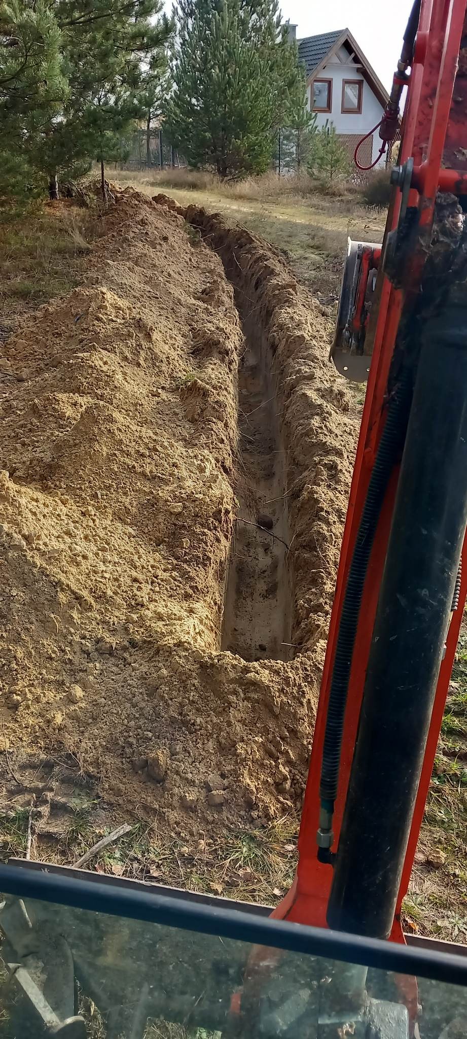 Wąski wykop w piaszczystej glebie, wykonany koparką; widok z kabiny operatora, w tle dom jednorodzinny.
