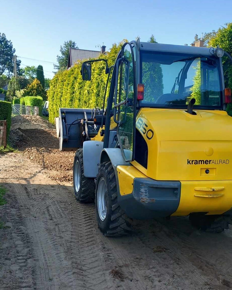 Żółta ładowarka Kramer Allrad 850 w trakcie pracy, widoczne ślady opon na piaszczystym podłożu, z tyłu usypisko ziemi i zielony żywopłot.