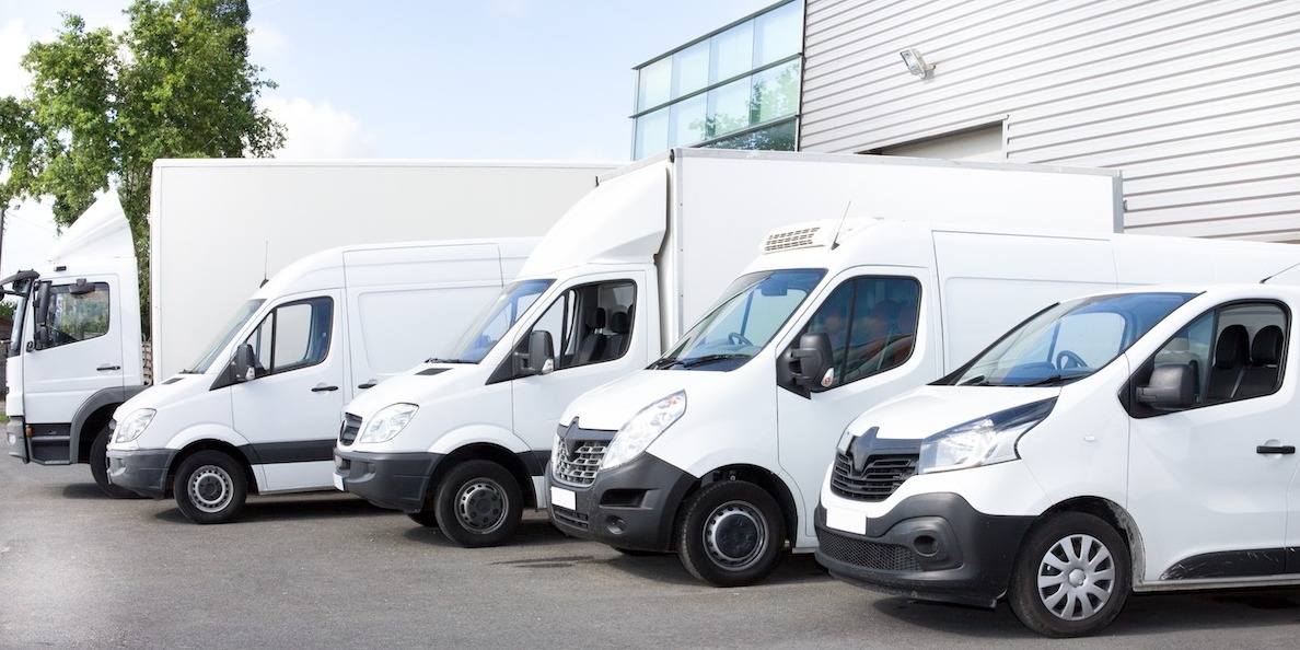 Rząd zaparkowanych białych samochodów dostawczych różnych typów, w tym ciężarówka z zabudową kontenerową, na tle budynku z szarymi panelami.