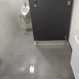 Wnętrze toalety publicznej z umywalką pisuarową, szarą podłogą z płytek i ciemnoszarymi drzwiami kabiny.