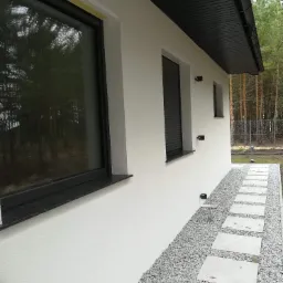 Nowoczesny dom jednorodzinny z białą elewacją, ciemną stolarką okienną i dachem, widok z boku na ścieżkę z płyt chodnikowych wysypaną żwirem.