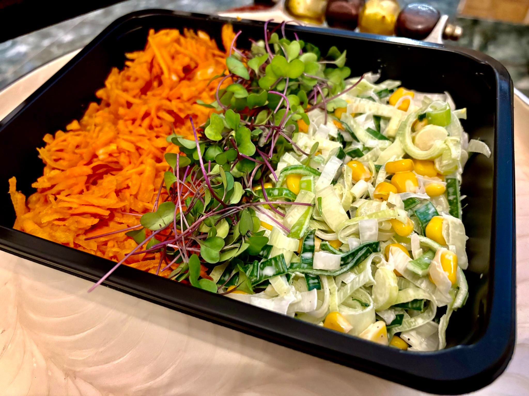 Gotowa sałatka w czarnym pojemniku: marchewka tarta, mikroliście i sałatka z pora z kukurydzą.