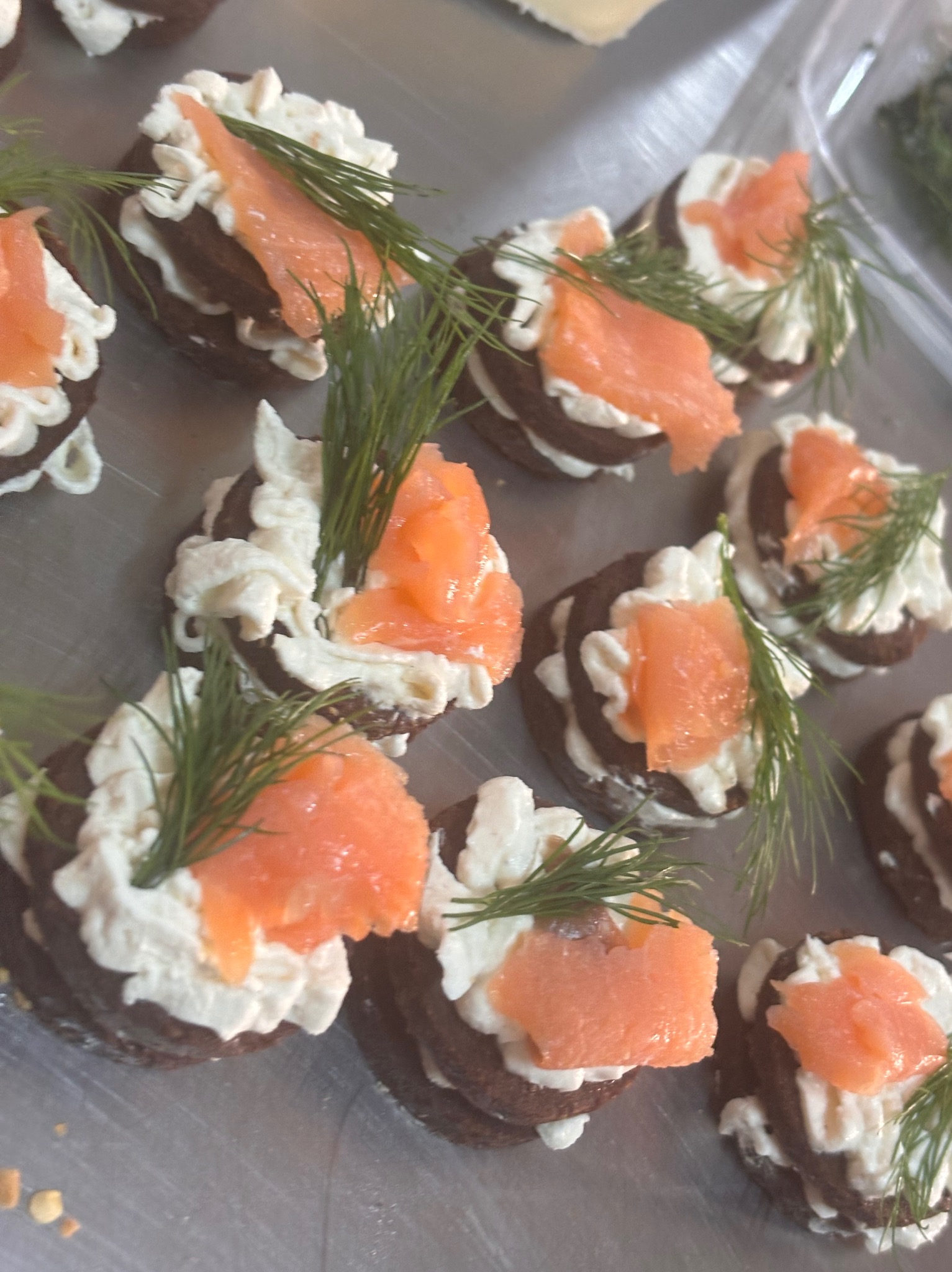 Mini kanapeczki z ciemnego pieczywa, kremowego serka, łososia i koperku, ułożone na metalowej tacy.