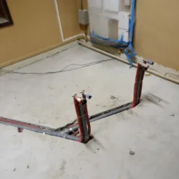 Szare rury instalacji wodnej i czerwone rury ogrzewania wystają z betonowej podłogi w trakcie remontu, zabezpieczony folią biały bojler w tle.