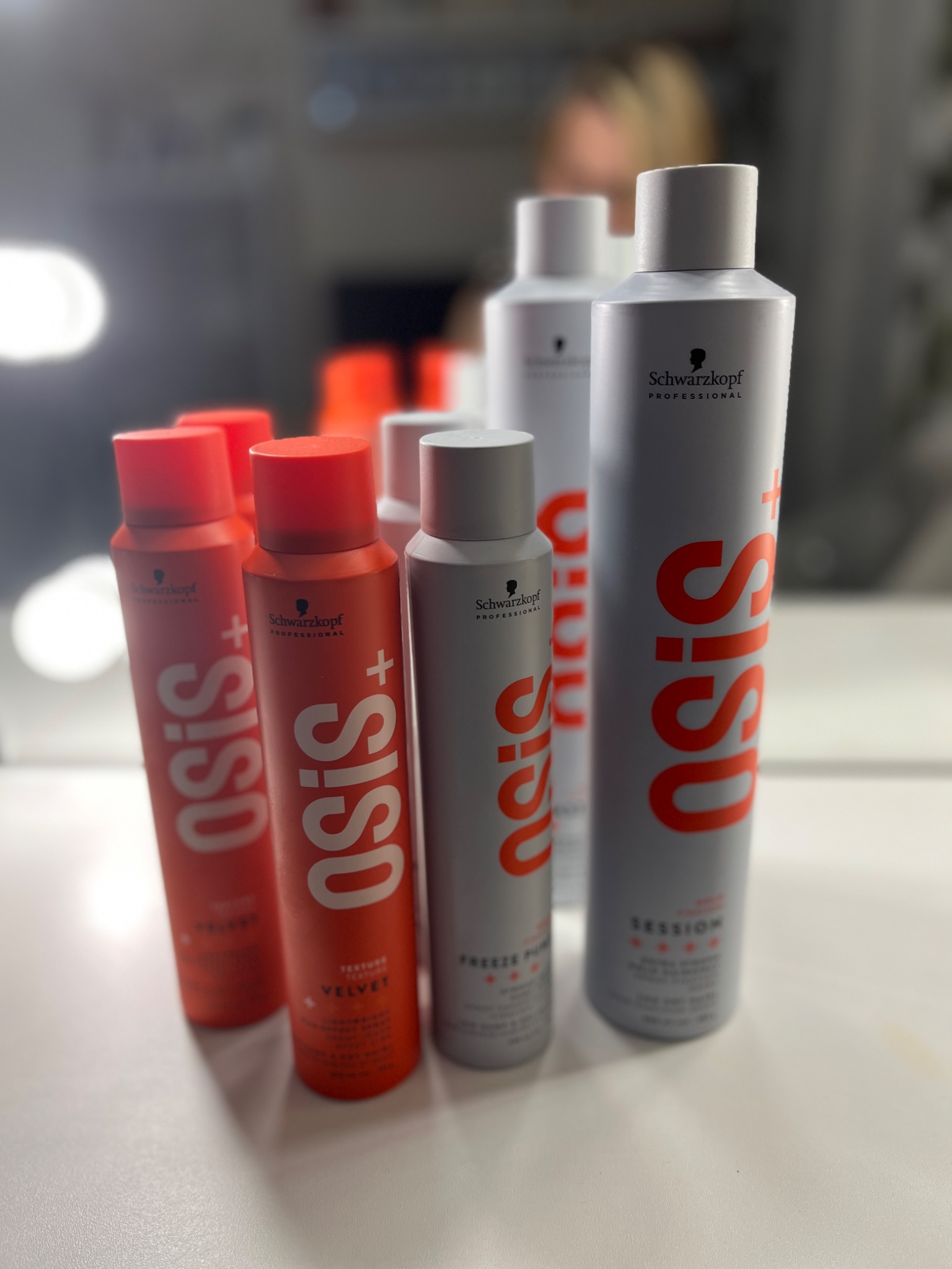 Różne produkty do stylizacji włosów marki Schwarzkopf Osis+ w odcieniach czerwieni i szarości, ustawione na białej powierzchni w salonie fryzjerskim, z rozmytym tłem lustra i oświetlenia.