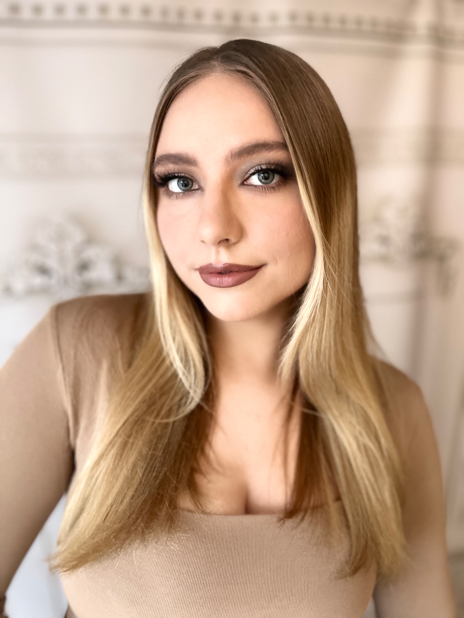 Portret kobiety z blond pasemkami, subtelnym makijażem oczu i ustami w odcieniu mauve, na tle kremowej ściany z delikatnym wzorem.