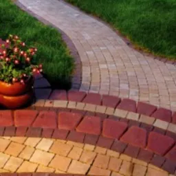 Kaskadowe schody z kostki brukowej w odcieniach czerwieni i beżu, prowadzące do krętej ścieżki z kostki w geometryczny wzór. Po lewej stronie donica z kwiatami i trawnik.