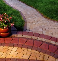Kaskadowe schody z kostki brukowej w odcieniach czerwieni i beżu, prowadzące do krętej ścieżki z kostki w geometryczny wzór. Po lewej stronie donica z kwiatami i trawnik.