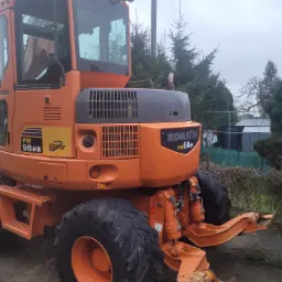 Pomarańczowa koparka kołowa Komatsu PW98MR zaparkowana w Szczecinie na nierównym terenie z widocznymi drzewami w tle.