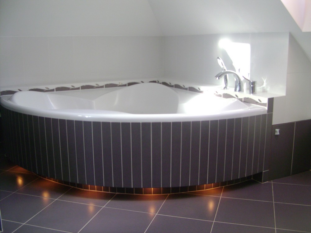 Nowoczesna, narożna wanna z hydromasażem, obudowana pionowymi panelami w odcieniach szarości z delikatnym podświetleniem LED u dołu, uzupełniona armaturą w kolorze chromu.