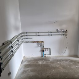 HYDRO-LUK USLUGI INSTALACYJNO-SANITARNE ŁUKASZ SZYBICKI - Świeżo zamontowane rury instalacji wodnej i kanalizacyjnej na ścianie w surowym wnętrzu, widoczny filtr AFRISO, instalator z tabletem w tle.