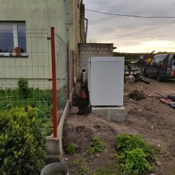 HYDRO-LUK USLUGI INSTALACYJNO-SANITARNE ŁUKASZ SZYBICKI - Biała pompa ciepła zainstalowana przy ścianie budynku, widoczne wykopy i instalacja, ogrodzenie z siatki w tle, pochmurne niebo.