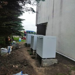 HYDRO-LUK USLUGI INSTALACYJNO-SANITARNE ŁUKASZ SZYBICKI - Trzy jednostki zewnętrzne, prawdopodobnie pomp ciepła, ustawione na betonowych podstawach przy ścianie budynku. Widoczny niezagospodarowany teren wokół, torba z grysem i pracownik w tle.