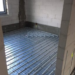 HYDRO-LUK USLUGI INSTALACYJNO-SANITARNE ŁUKASZ SZYBICKI - Białe rury ogrzewania podłogowego ułożone na czarnej siatce w surowym pomieszczeniu z białych bloczków, widoczne okno i ślady po instalacjach na ścianach.