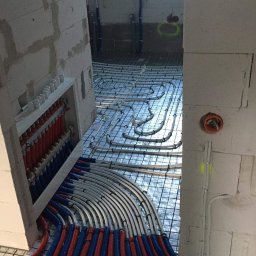 HYDRO-LUK USLUGI INSTALACYJNO-SANITARNE ŁUKASZ SZYBICKI - Instalacja ogrzewania podłogowego w surowym wnętrzu, rury grzewcze ułożone na siatce, widoczny rozdzielacz z rurami w kolorze czerwonym i niebieskim, ściany z betonu komórkowego.