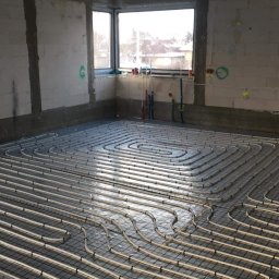 HYDRO-LUK USLUGI INSTALACYJNO-SANITARNE ŁUKASZ SZYBICKI - Wnętrze w trakcie montażu ogrzewania podłogowego: rury ułożone w spiralny wzór na siatce, surowe ściany z betonu komórkowego, okno z widokiem na zabudowę.