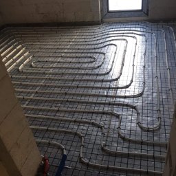 HYDRO-LUK USLUGI INSTALACYJNO-SANITARNE ŁUKASZ SZYBICKI - Układanie rur ogrzewania podłogowego na siatce montażowej, widoczne fragmenty rur doprowadzających w kolorze niebieskim i czerwonym, surowe ściany z betonu komórkowego.