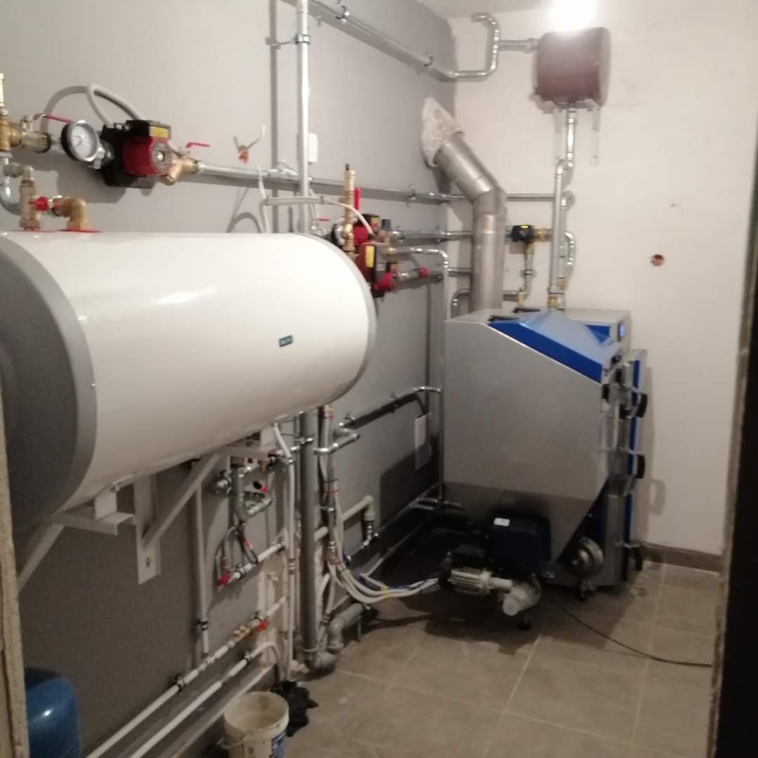 Kompletna instalacja kotłowni z kotłem na paliwo stałe, zasobnikiem ciepłej wody i rozbudowaną siecią rur, zaworów oraz osprzętu, widoczne elementy montażu.