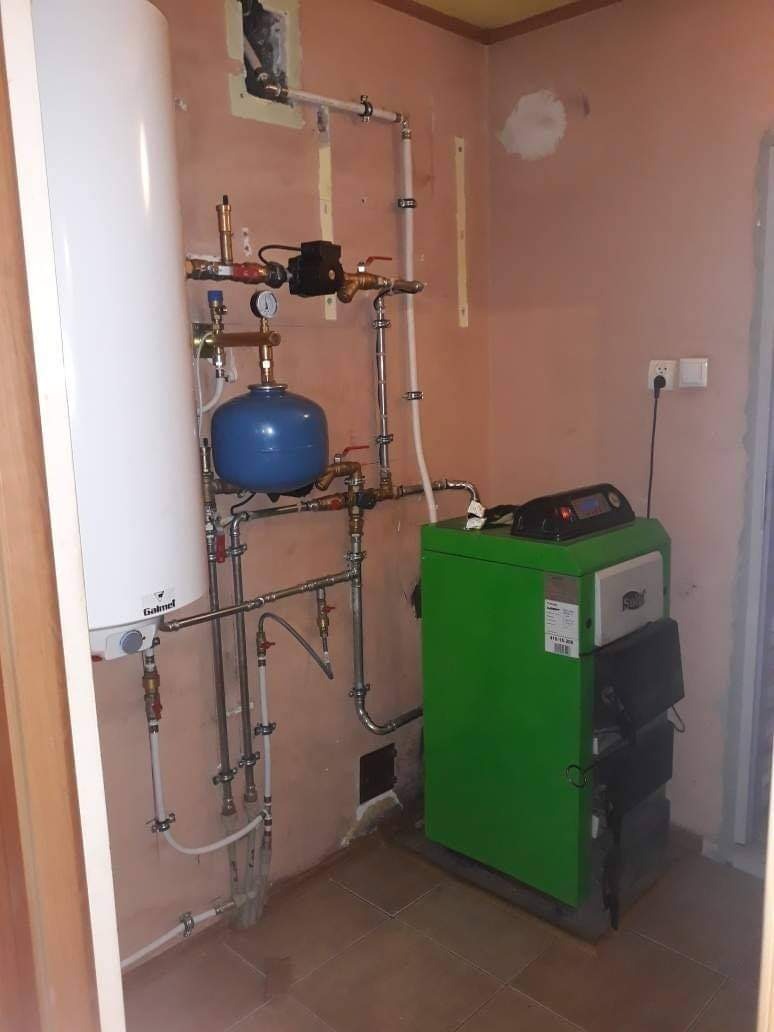 Zainstalowany, zielony piec centralnego ogrzewania z widocznymi rurami, zbiornikiem wyrównawczym i osprzętem, na tle pomalowanej ściany w trakcie remontu.