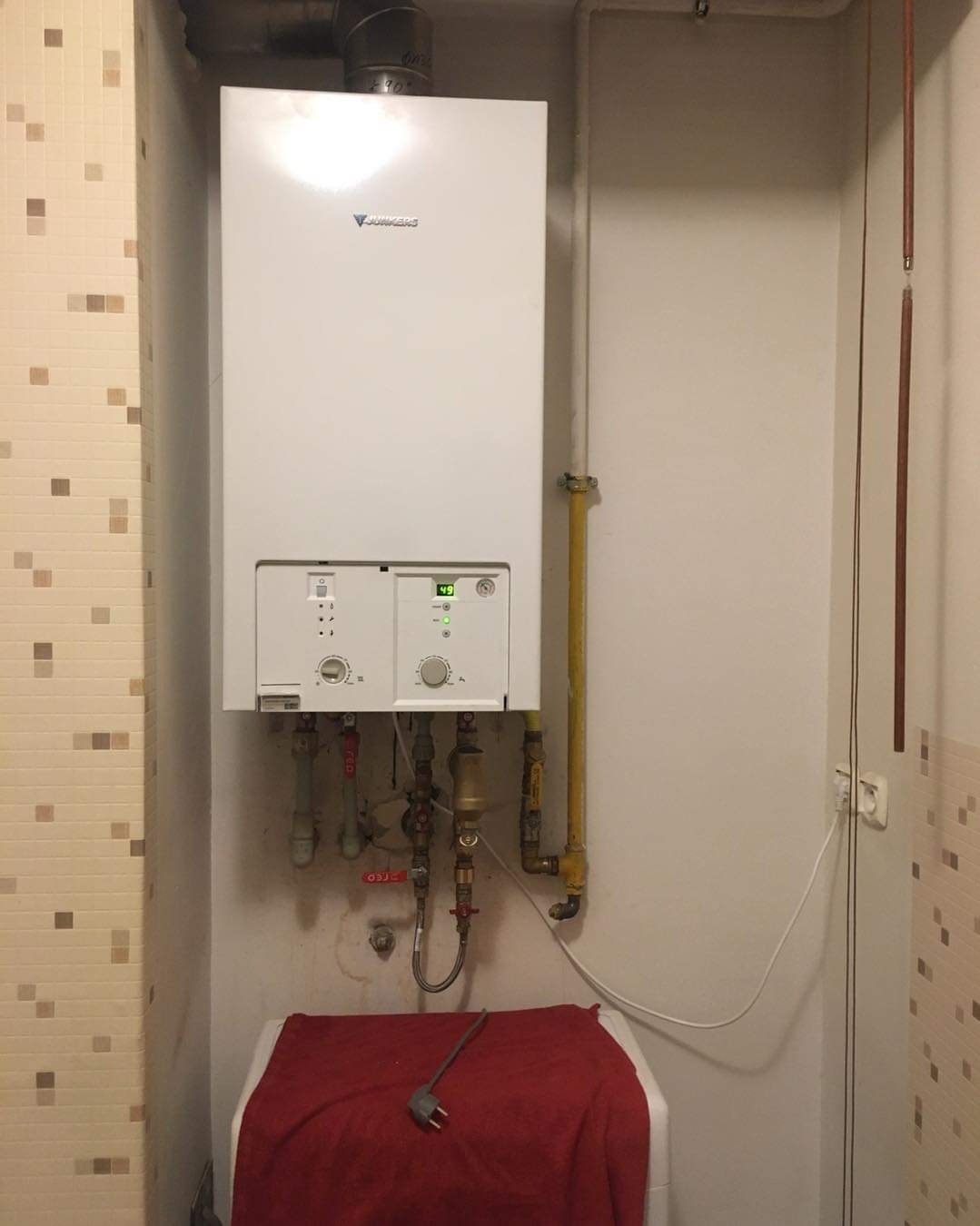 Biały piec gazowy Junkers zamontowany na ścianie w małej kotłowni, widoczne rury gazowe, instalacja wodna i czerwony ręcznik na pralce pod spodem.