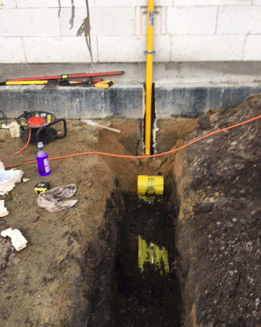 Wykop z żółtą rurą gazową pionowo wzdłuż ściany fundamentowej, widoczna pomarańczowa rura, narzędzia i butelka w tle.