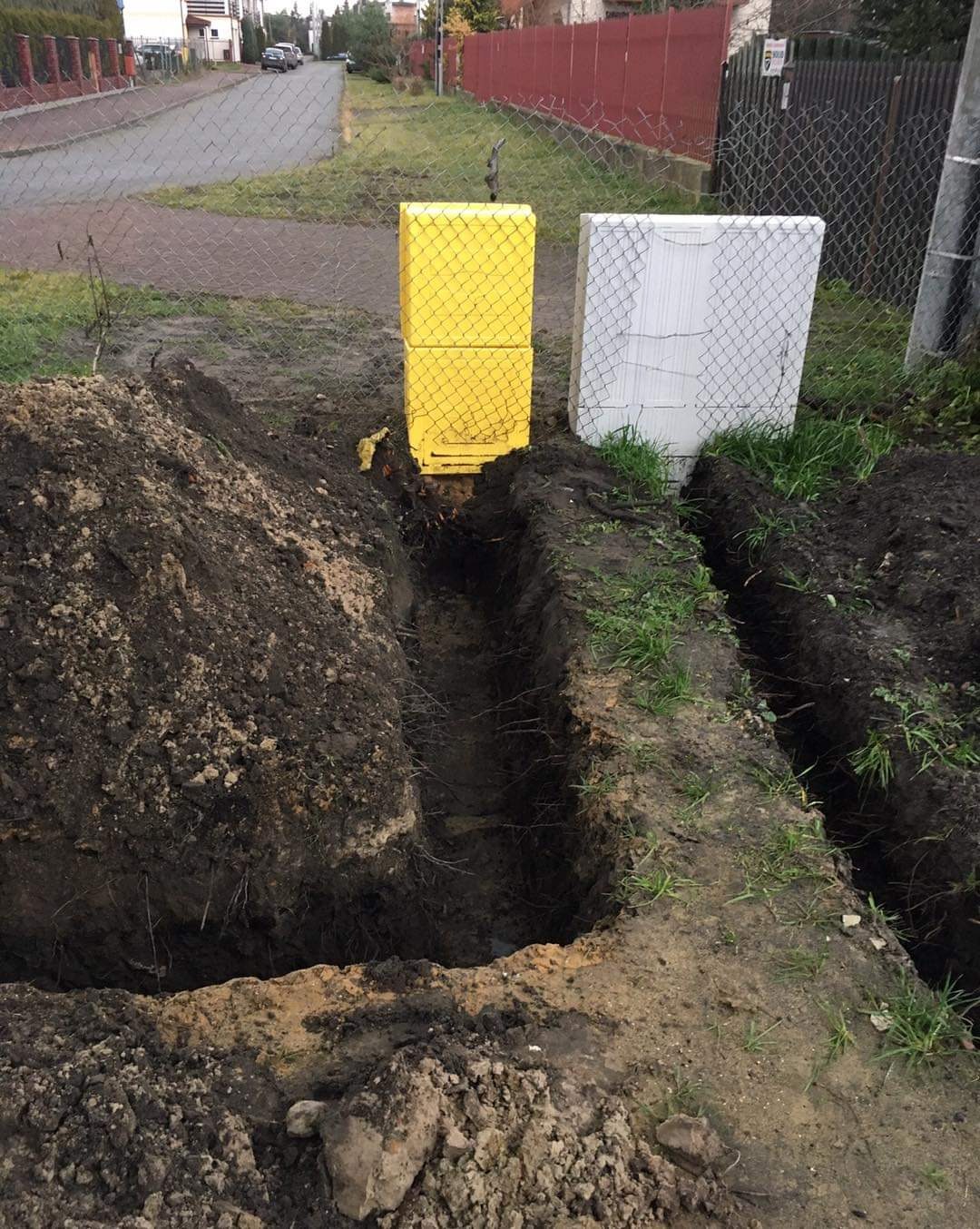 Wykonane wykopy pod instalację dwóch skrzynek, jedna żółta, druga biała, na zewnątrz budynku, widok z poziomu gruntu.