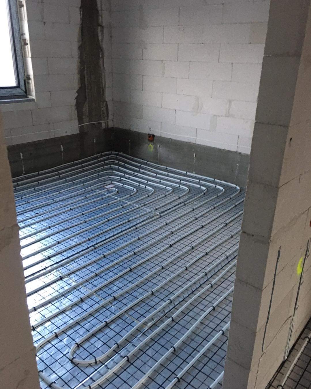 Białe rury ogrzewania podłogowego ułożone na czarnej siatce w surowym pomieszczeniu z białych bloczków, widoczne okno i ślady po instalacjach na ścianach.