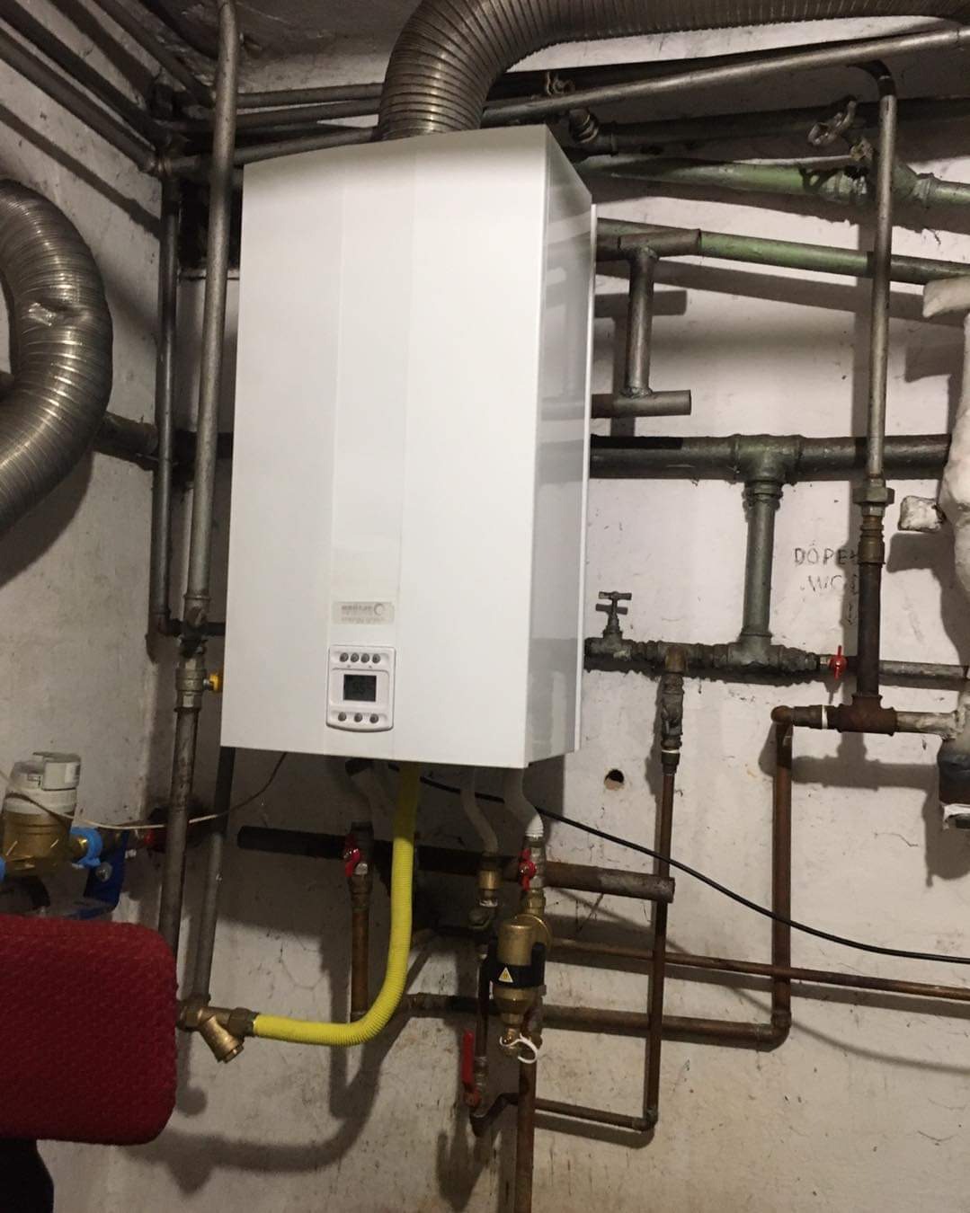 Nowy, biały piec gazowy zamontowany na tle starej, pomalowanej ściany z widocznymi rurami i przyłączami gazowymi. Żółty, elastyczny przewód gazowy łączy piec z instalacją.