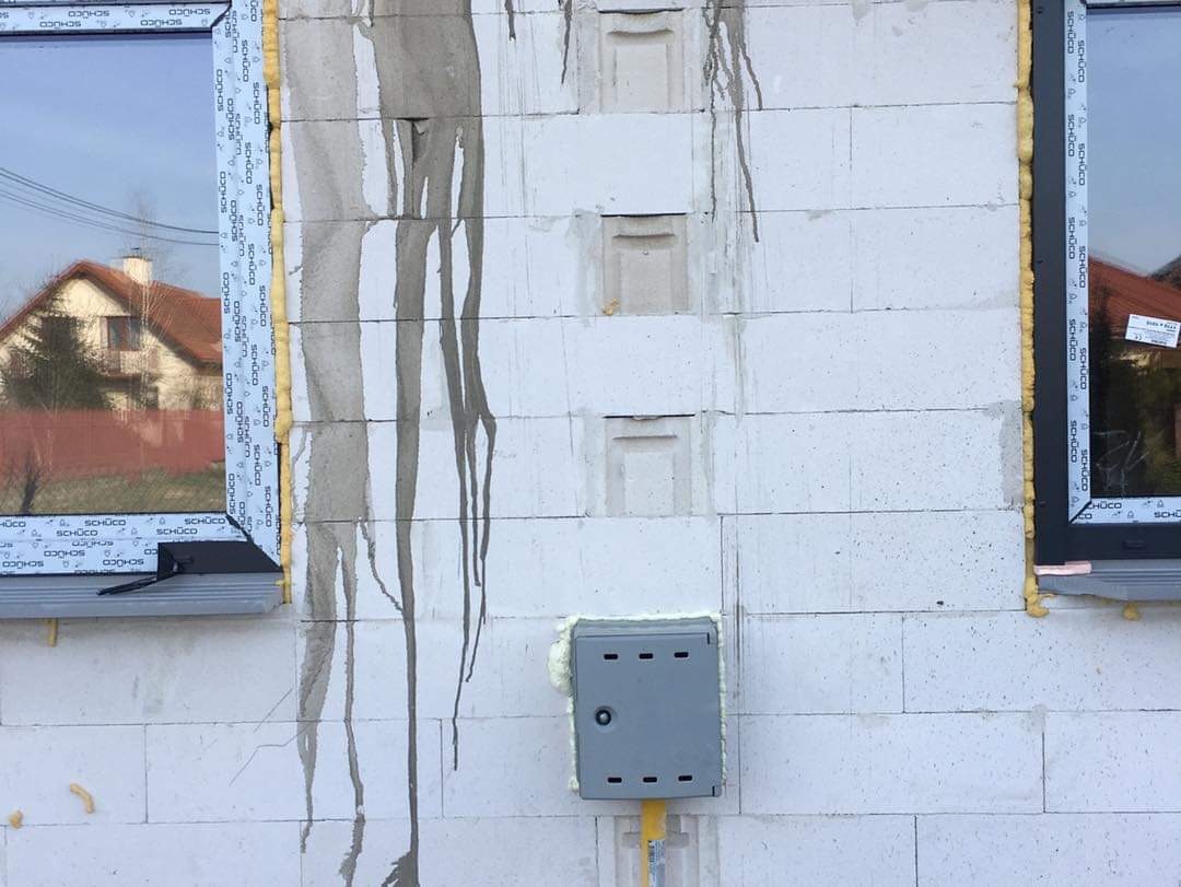 Szara skrzynka gazowa z żółtą rurą na ścianie z betonu komórkowego, widoczne ślady zaprawy, okna z ramami Schüco w tle.