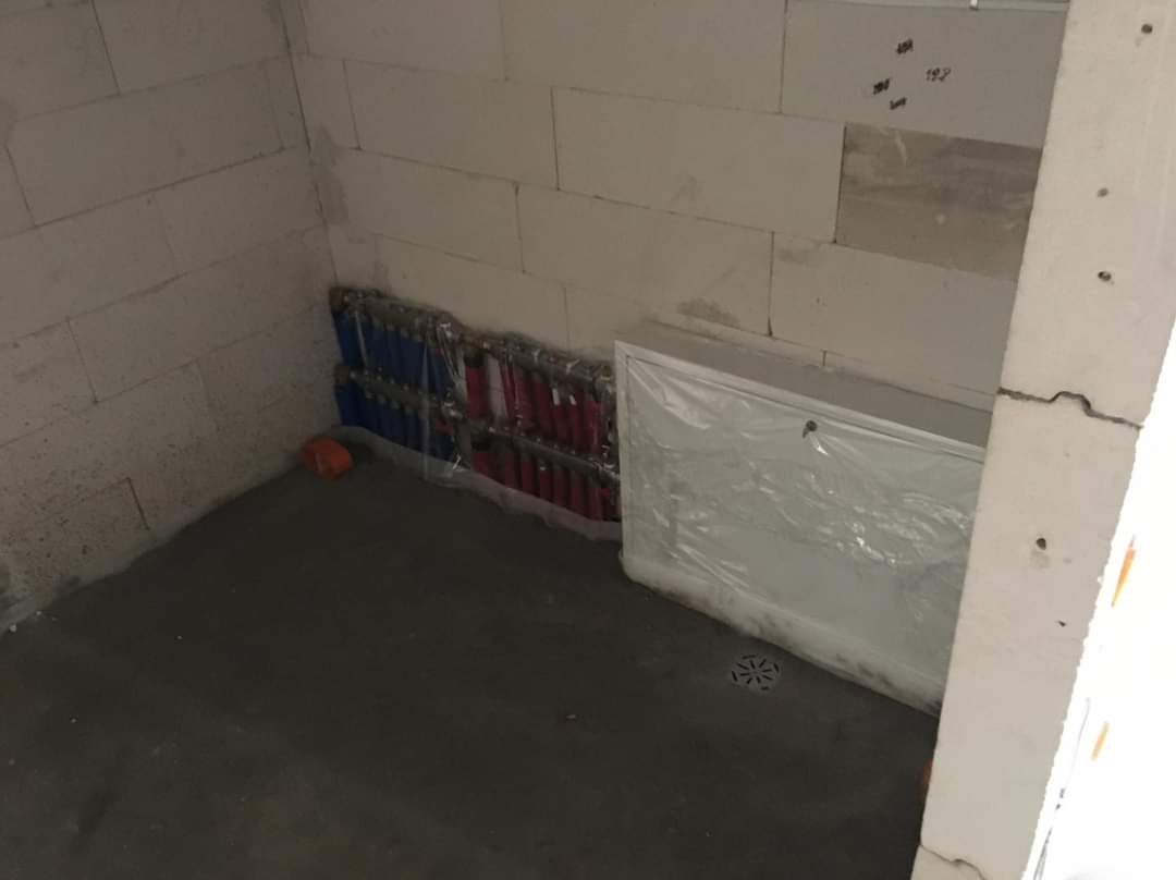 Surowy stan instalacji podłogowej w pomieszczeniu z betonową wylewką, widoczne rury grzewcze w kolorze czerwonym i niebieskim oraz odpływ liniowy zabezpieczony folią.