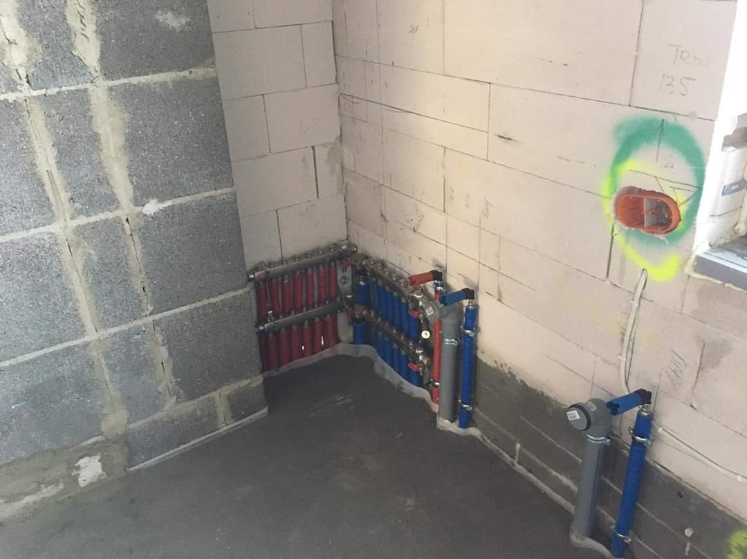 Rozdzielacz instalacji centralnego ogrzewania z rurami w kolorze czerwonym i niebieskim, zamontowany na ścianie z betonu komórkowego w surowym pomieszczeniu. Widoczne także rury doprowadzające wodę...