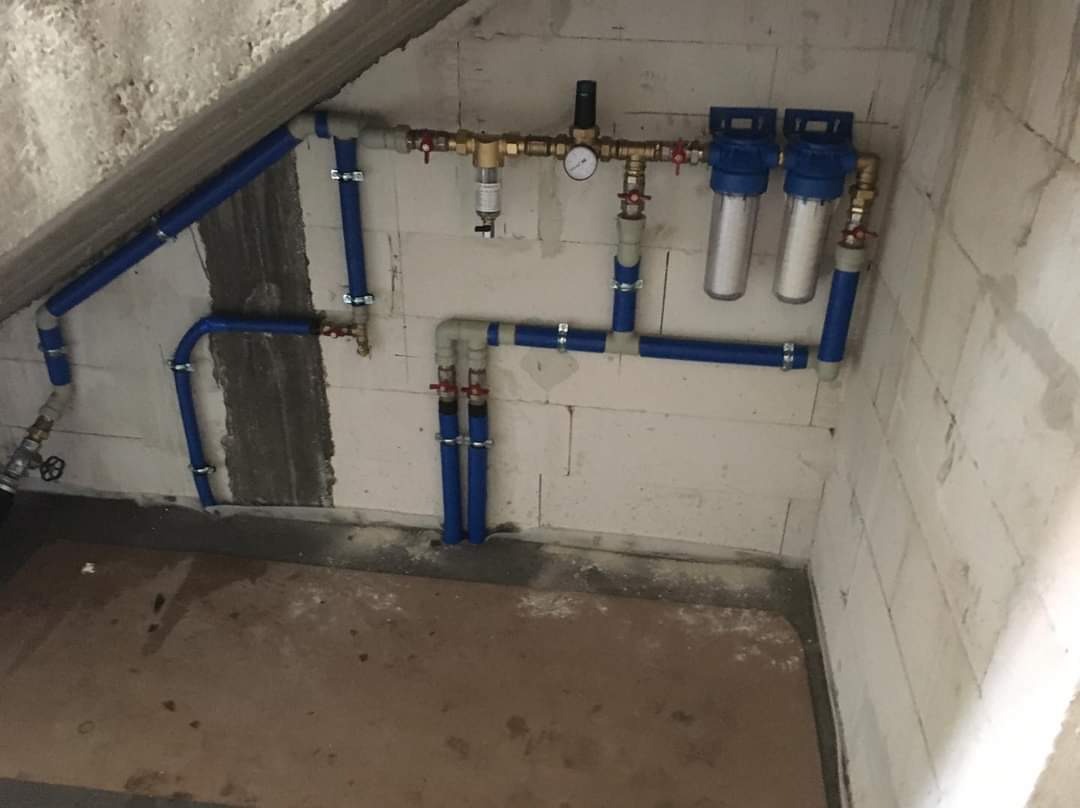 Instalacja filtrów wody z manometrem i zaworami, rury w kolorze niebieskim, zamontowane na ścianie z betonu komórkowego w surowym stanie.