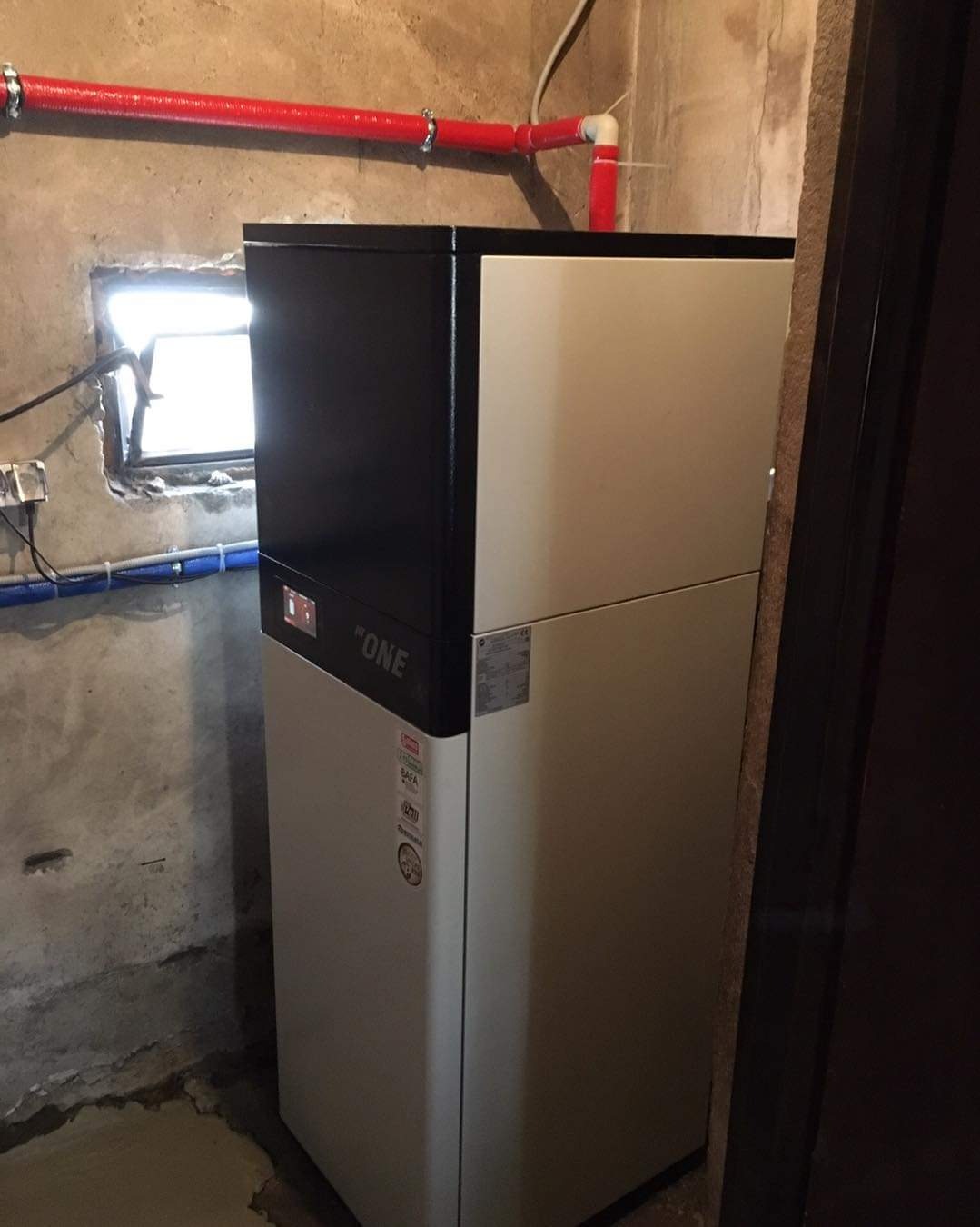 Kompaktowa, dwuczęściowa pompa ciepła w piwnicy z małym oknem i widocznymi rurami instalacji. Model oznaczony napisem 'ONE'.
