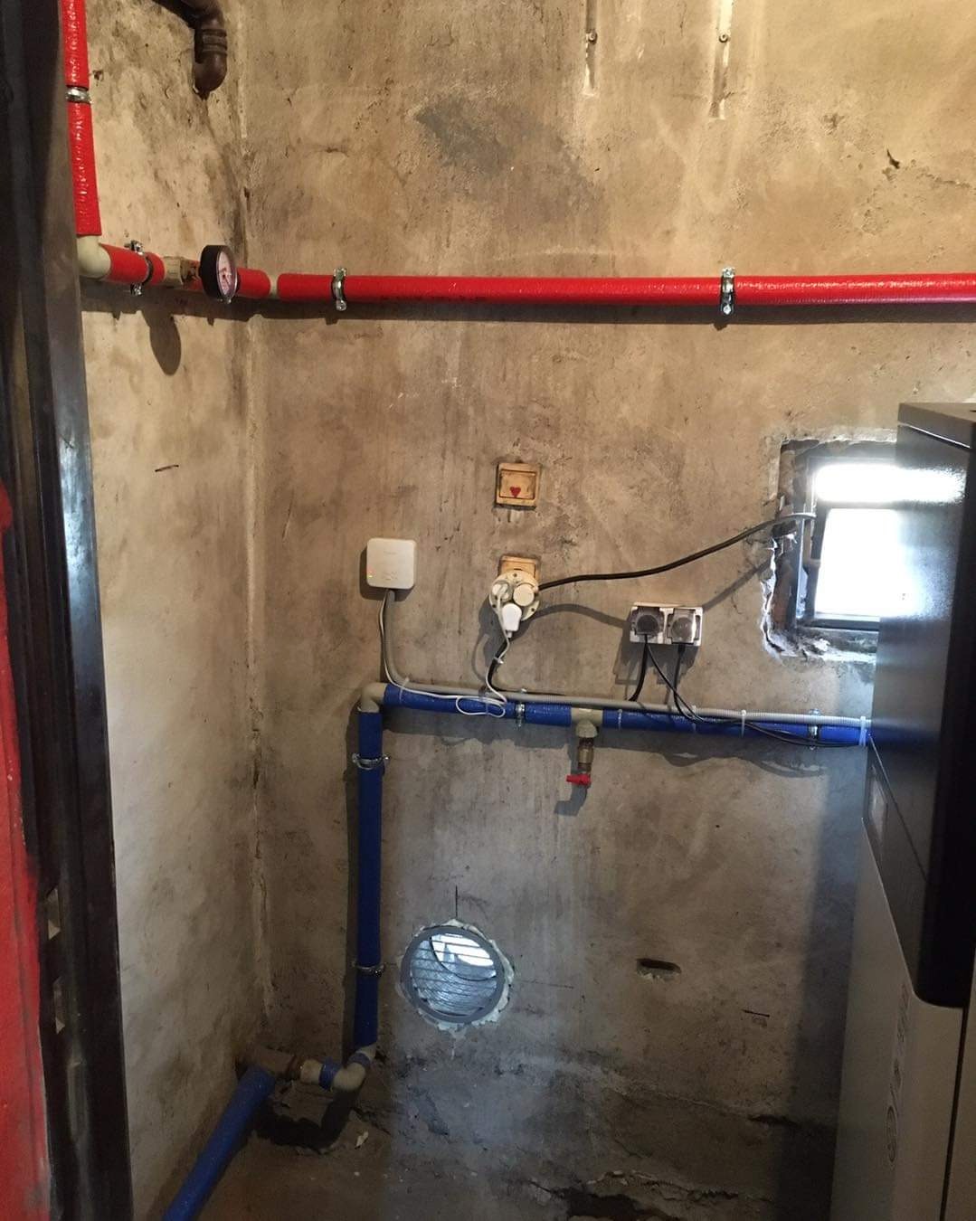 Fragment kotłowni z widocznymi instalacjami: czerwone rury izolowane termicznie pod sufitem z manometrem, niebieskie rury z zaworem, gniazdka elektryczne na szorstkiej ścianie oraz okienko w rogu.