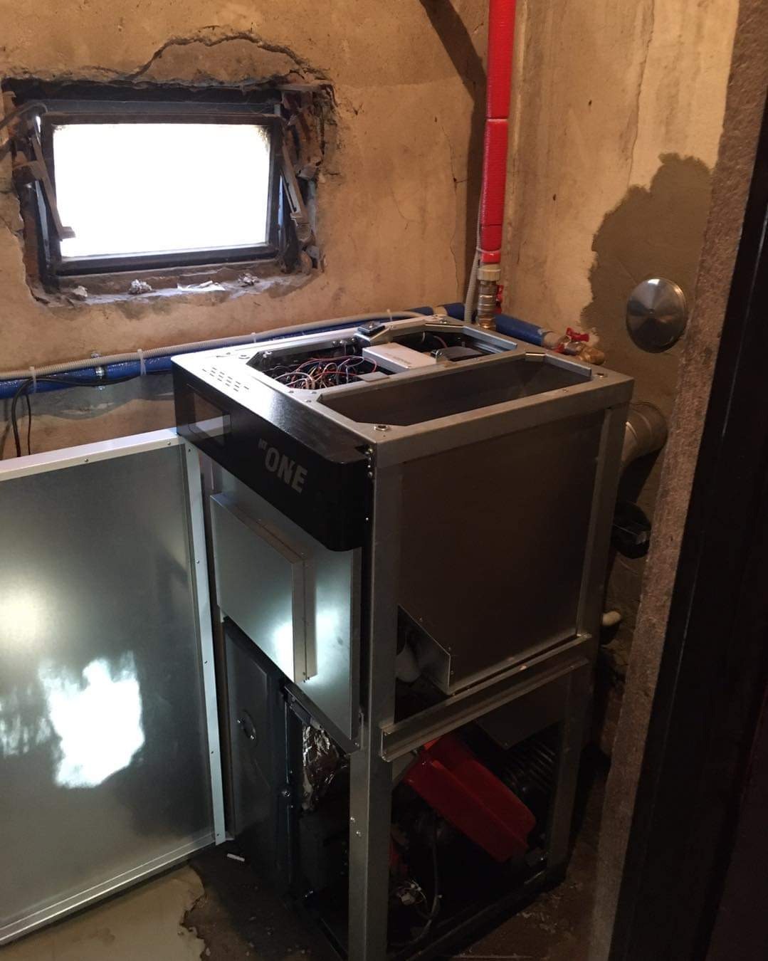 Otwarta obudowa pompy ciepła 'ONE' w surowym wnętrzu z oknem i widocznymi instalacjami hydraulicznymi