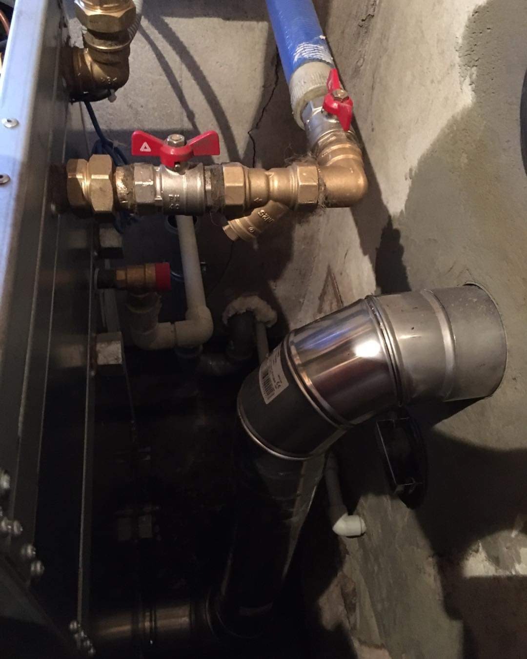Szczegółowe ujęcie instalacji hydraulicznej z zaworami, rurami miedzianymi i plastikowymi, oraz fragmentem metalowego przewodu wentylacyjnego w surowym, betonowym otoczeniu.