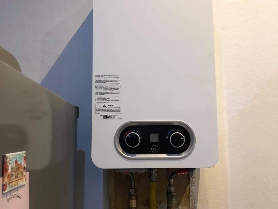 Biały, wiszący piec gazowy z czarnym panelem sterowania, zamontowany na ścianie z widocznymi rurami doprowadzającymi gaz, obok lodówki z magnesem 'Wrocław'.