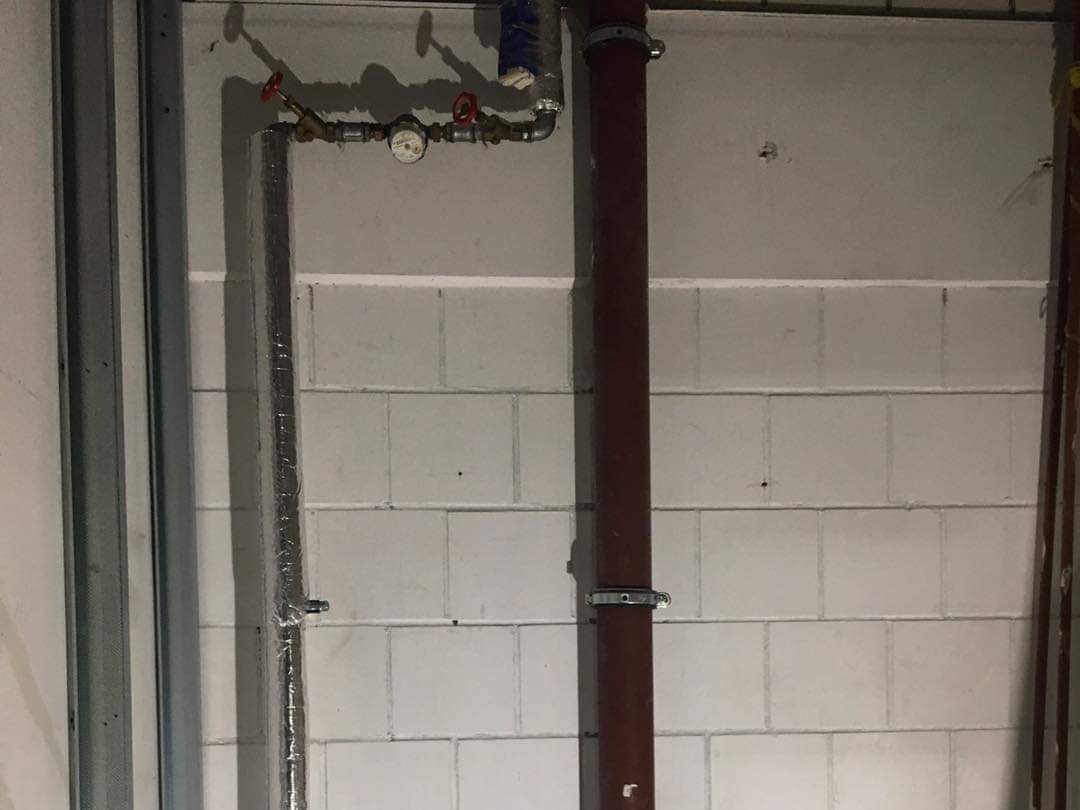 Rury instalacyjne z manometrem i zaworami zamontowane na ścianie z bloczków betonowych, widoczne elementy konstrukcji szkieletowej z metalu.