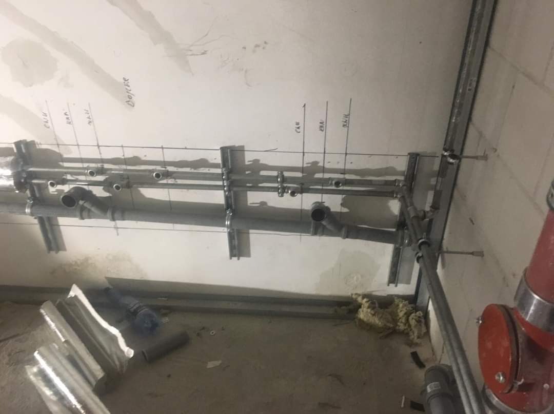 Montaż instalacji hydraulicznej pod sufitem, widoczne rury, profile montażowe, oznaczenia na ścianie i fragment czerwonej instalacji przeciwpożarowej.